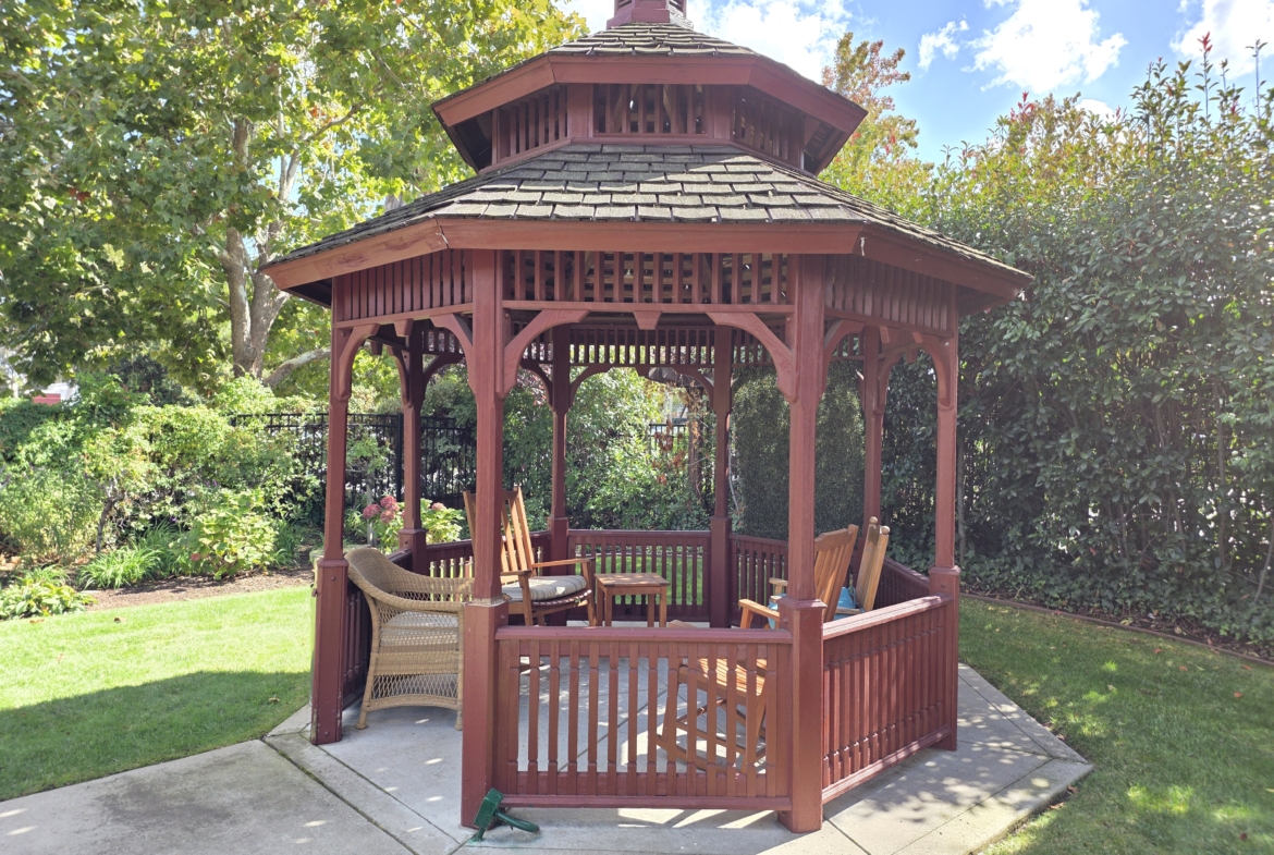 Wooden gazebo with chairs in garden setting.