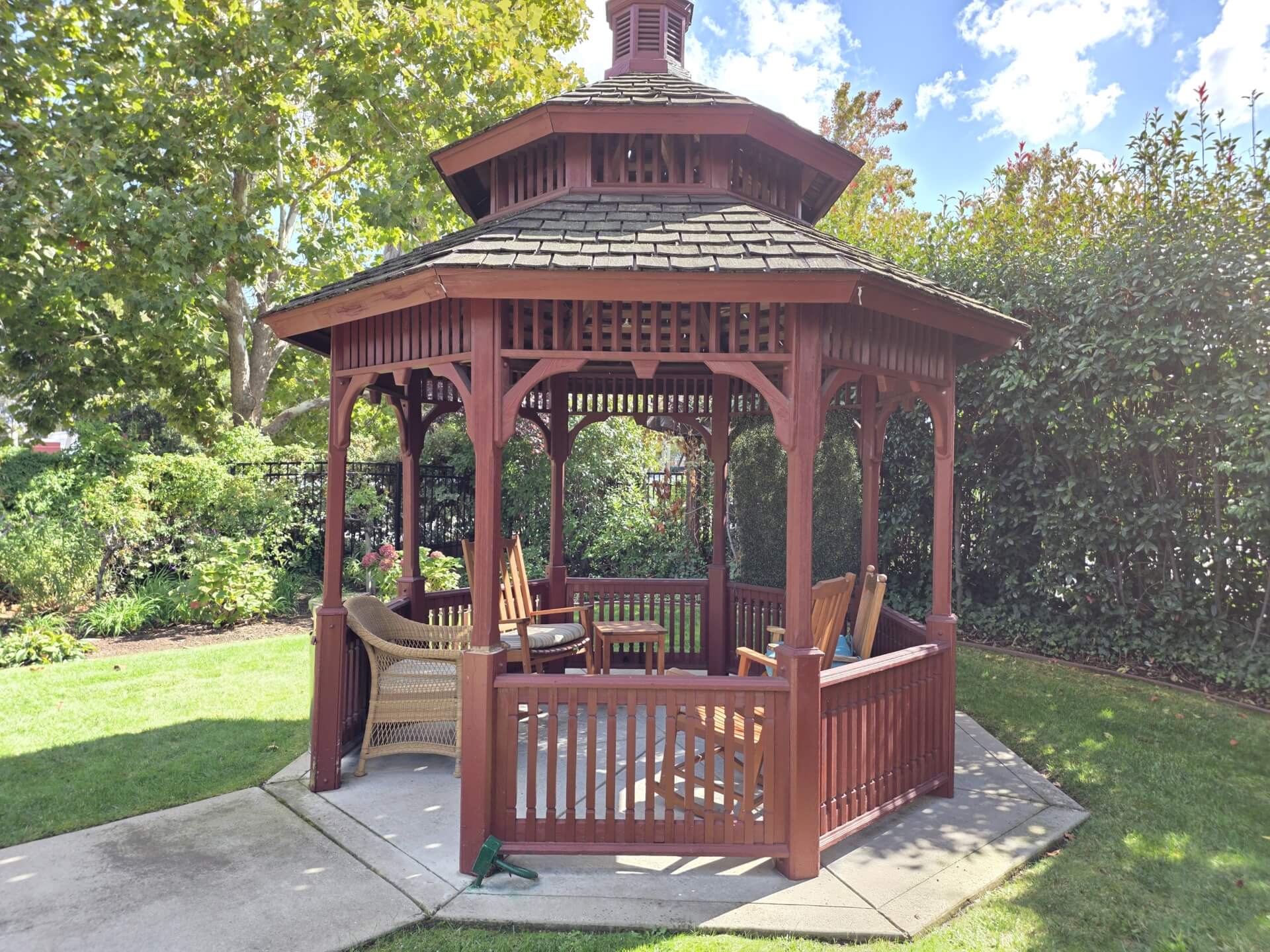 Wooden gazebo with chairs in garden setting.